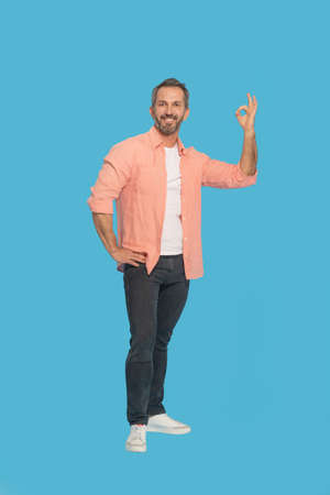 Gesturing OK handsome middle aged grey haired man happy smiling looking at camera wearing peach shirt isolated on blue background. Fit mature man saying im OK.の写真素材