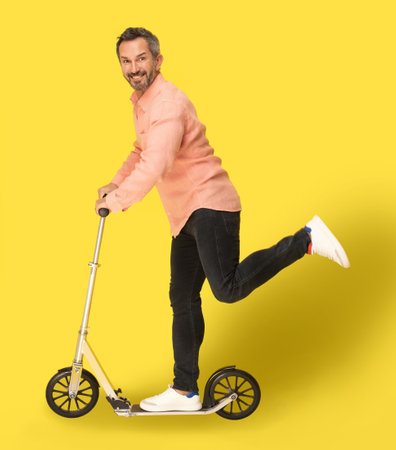 Grey haired middle aged man ride scooter stretched leg up looking at camera wearing peach shirt and black jeans isolated on yellow background. Handsome mature businessman on the go.の写真素材