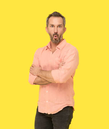 Amazed facial expression middle aged grey haired man standing with arms folded happy looking at camera wearing pink shirt isolated on yellow background. Mature fit man, healthy lifestyle concept.の写真素材