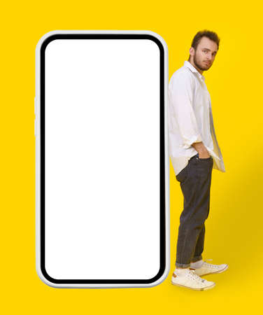 Young man leaned on big smartphone in casual wear and eye glasses posing on yellow background. Stylish bearded smart casual look hipster man with phone. Mobile app advertisement mock up.の写真素材