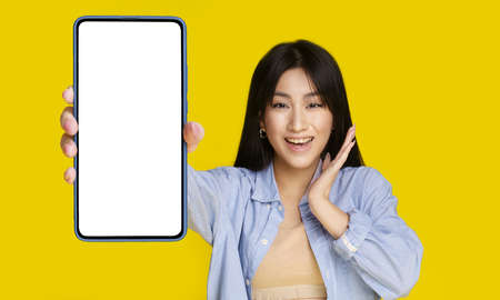 Beautiful asian girl wowing facial expression holding smartphone with white screen happy to introduce new app, game, win, isolated on yellow background. Product placement. Mobile app advertisement.の写真素材