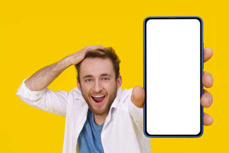 Handsome excited young man emotionally smiling touching his head showing huge smartphone with white screen. Mobile app advertising mockup product placement. Young man with phone.の写真素材