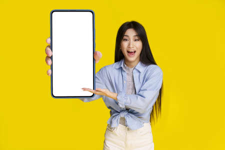 Big smartphone with white screen in hand of beautiful asian girl happy to introduce new app, game, win, isolated on yellow background. Mobile app advertisement product placement.の写真素材