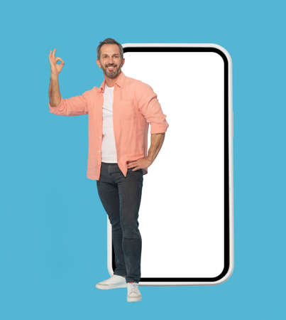 Gesturing OK standing in front big smartphone with white screen handsome middle aged grey haired man...