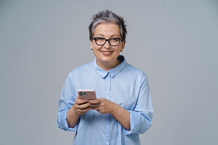 Texting or typing holding smartphone in hands mature grey haired woman working or shopping online, checking on social media. Pretty woman in blue blouse isolated on white background.の写真素材