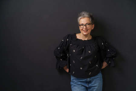 Gorgeous mature grey haired businesswoman with grey hair leaned on wall isolated on black background. Elegant woman with short hair. Image of successful mature woman. Aged beauty.の写真素材