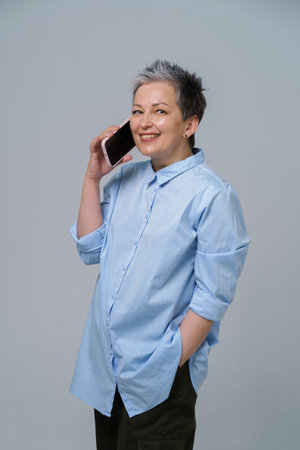 Mature woman with grey hair talking holding smartphone at her ear looking sideways, smiling wearing long blue shirt. Pretty woman in blue blouse isolated on white background.の写真素材