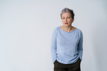 Stressed, angry mature grey haired woman 50s posing frustrated looking at camera with hands in pockets, copy space on left isolated on white background. Mature people healthcare.の写真素材