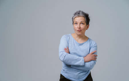 Grey haired mature woman standing with arms folded and wondering face expression in blue blouse copy space on left isolated on white background. Healthcare concept. Aged beauty concept. Copy space.の写真素材