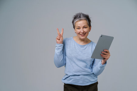 Gesturing V or victory mature grey haired woman in 50s holding digital tablet working or shopping online, checking on social media. Pretty woman in blue blouse isolated on white background.の写真素材