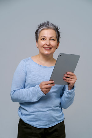 Mature grey haired woman holding digital tablet working or shopping online, checking on social media. Pretty woman in blue blouse isolated on white background.の写真素材
