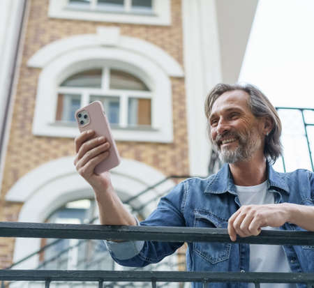 Happy middle aged grey bearded man laughing reading text message or funny social media post using smartphone standing in old town wearing casual. Travel concept.の写真素材