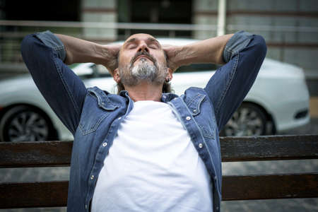 Relaxed, enjoying life handsome middle age man sits leaned back with hands behind his head on the bench outdoors at urban city background wearing denim shirt and white t-shirt. Travel concept.の写真素材