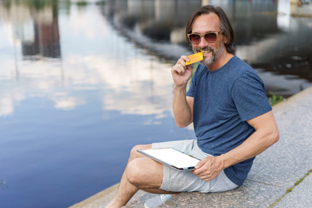 Biting credit, debit card excited freelancer man sitting outdoors next to river working using digital tablet pc, shopping online or paying online banking. E-banking concept. Financial concept.の写真素材