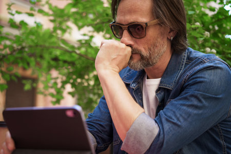 Puzzled up, sad middle aged man working outdoors using digital tablet. Handsome freelancer traveling man not happy checking work or social media sitting at city street cafe.の写真素材
