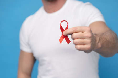 Man with red ribbon bow AIDS awareness symbol in hand wearing a white t-shirt isolated on a blue background. Modern medicine and healthcare. AIDS awareness concept. No face visible.の写真素材