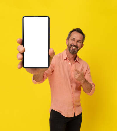 Happy middle aged grey haired fit man holding big smartphone pointing finger at it wearing peach shirt isolated on yellow. Mature muscled man with phone app advertisement. Mock up white screen.の写真素材