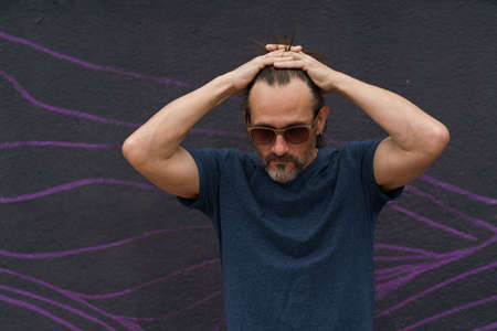 Thoughtful mature man standing with hand folded above his head next to dark urban wall leaned head...