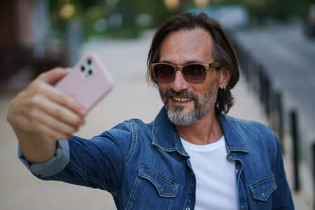 Man with smartphone outdoors making selfie or recording video message wearing denim shirt and sunglasses. Handsome middle aged man with grey hair making photos while traveling.の写真素材