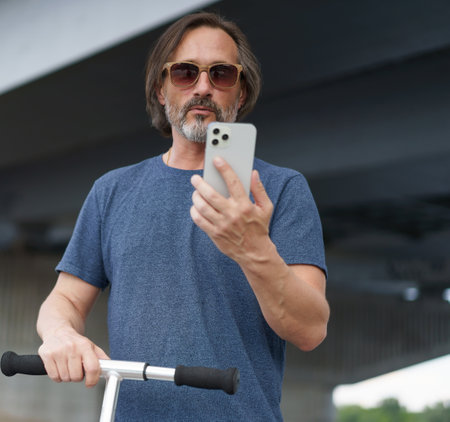 Mature man looking at phone in city. Man made stop using scooter answer the phone. Travel concept. Handsome stylish sexy middle aged grey bearded man in sunglasses under the bridge wearing casual.の写真素材