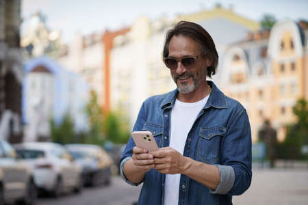 Texting or posting in social media post, photo mature handsome man with smartphone outdoors wearing...
