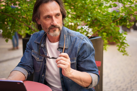 Middle age freelancer businessman working outdoors sitting at street cafe using digital tablet planing project. Middle aged man looking away sitting at street restaurant during vacation traveling.の写真素材