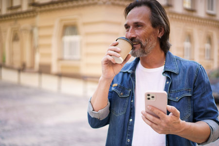 Mature man with coffee and smartphone outdoors. Middle aged man read having video call holding phone and take away paper cup with coffee wearing denim shirt. Business on the go concept.の写真素材