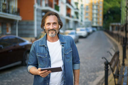 Middle aged grey haired freelancer man hold digital tablet in hands having a call while standing on urban streets. Mature man talking or listening music use wireless earphones travel old town.の写真素材