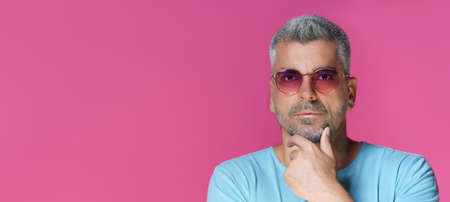 Thoughtful handsome man 30s 40s wearing pink sunglasses and blue t-shirt touching his chin with pensive, concentrated look isolated on pink background. Grey haired mature man studio shot.の写真素材
