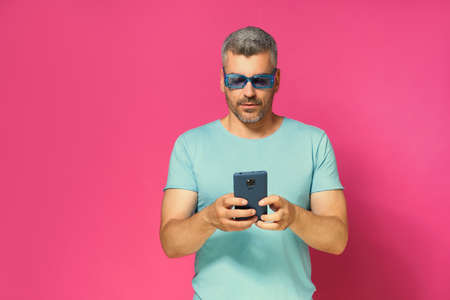 Concentrated, pensive handsome man 30s 40s looking down at smartphone in hands wearing blue sunglasses and t-shirt isolated on pink background. Mucho man in blue t-shirt with phone studio shot.の写真素材