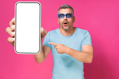 Shocked handsome man 40s pointing at big smartphone in hand wearing blue shirt and sunglasses isolated on pink background. Man with phone studio shot. Mobile app advertisement. Copy space.の写真素材