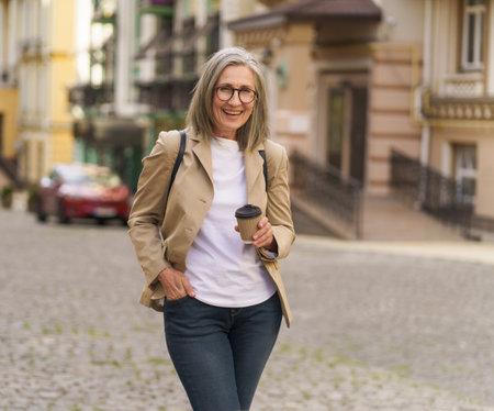 Excited mature businesswoman in casual jacket and denim jeans hold paper cup with hot coffee standing outdoors of old town. Senior woman spend traveling in European city enjoying cup of tea, coffee.の写真素材