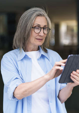 Interested in reading news or social media mature grey hair woman holding digital tablet sitting standing outdoors at the city streets. Mature woman answering call outdoors.の写真素材
