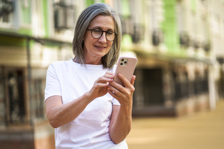 Mature woman with modern technologies. Senior grey hair lady holding smartphone standing outdoor at the streets of old town. Mature woman read text message standing or walking outdoors.の写真素材