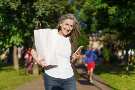 Charming mature woman walking in streets holding grocery paper bag with food and smartphone, phone in other hand cheerful smiling with blurred people life on background.の写真素材