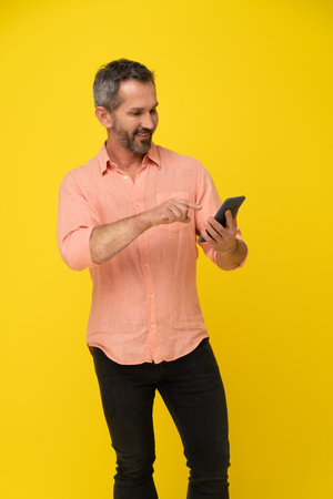 Middle aged grey haired handsome fit man holding smartphone looking at it wearing peach shirt isolated on yellow. Mature muscled man with phone, app advertisement.の写真素材