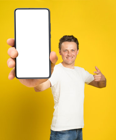 Holding huge smartphone with white screen handsome man pointing at white empty screen, wearing white t-shirt and jeans isolated on yellow background. Mobile app advertisement, great offer.の写真素材