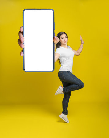 Charming asian middle age woman jump showing smartphone with white screen wearing white t-shirt and denim jeans, mobile app advertisement isolated on yellow background.の写真素材