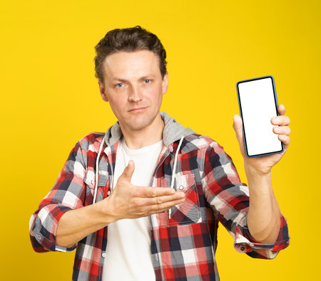 New app or website introduction holding smartphone with white screen blond man, wearing red plaid shirt. Man with phone display mock up isolated on yellow background. Mobile app advertisement.の写真素材