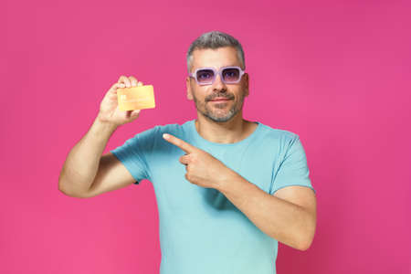 Serious handsome middle aged man hold debit, credit card in hand pointing at it wearing blue t-shirt and glasses isolated on pink background. Financial, banking concept.の写真素材
