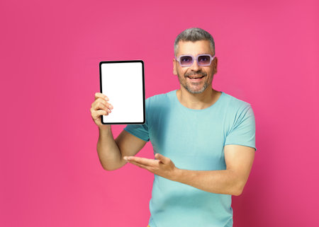 Introducing great offer showing digital tablet white screen handsome man looking at camera wearing casual blue shirt and sunglasses isolated on pink background. App advertisement.の写真素材