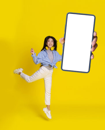 Mobile app great offer, jumping asian girl with big smartphone in hand in casual, isolated on yellow background. Full length portrait of joyful asian girl. Product placement.の写真素材
