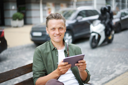 Portrait of young freelancer man work using digital tablet sits on the bench outdoors in front office building wearing casual green shirt and shoulder bag. Young handsome man working on the go.の写真素材