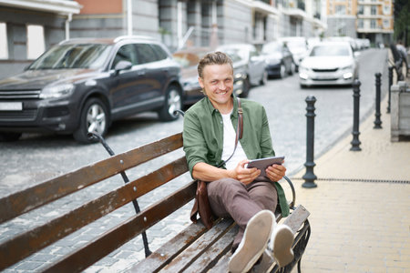 Happy freelancer man work using digital tablet sits on the bench outdoors in front office building wearing casual green shirt and shoulder bag. Young handsome man working on the go.の写真素材