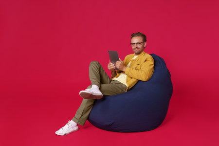 Handsome full size body young man 20s, 30s wearing casual denim clothes sit in bag chair holding digital tablet in hands working or looking at social media isolated on red background. Freelancer man.の写真素材