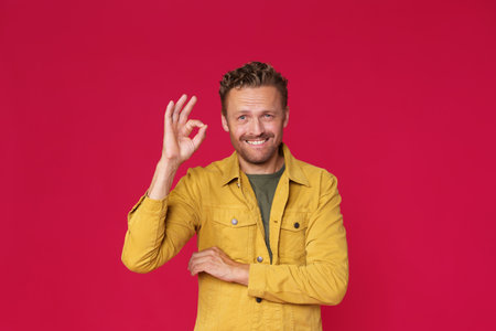 Cheerful young man showing, gesturing OK, okay smiling looking at camera wearing casual jeans yellow jacket isolated on red background. Happy handsome caucasian man in casual.の写真素材