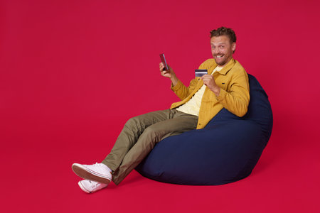 Buying online using debit, credit card handsome young man 30s wearing casual denim clothes sit in bag chair holding digital tablet in hands working isolated on red background. Freelancer man.の写真素材