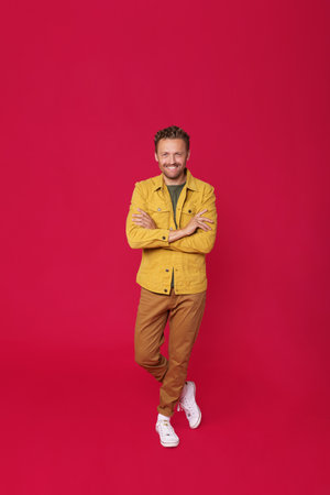 Smiling handsome young man in jeans clothes and white running shoes standing with arms and feet crossed isolated on red background. Happy freelancer man enjoy his success project.の写真素材