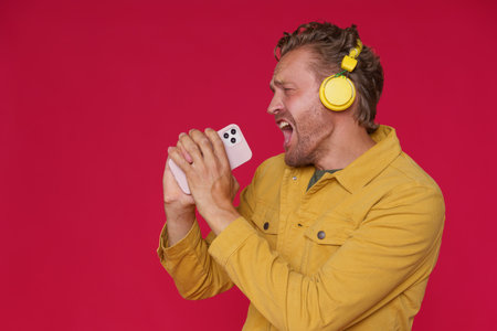 Handsome man 30s singing enjoying his favorite song or track using phone and wireless headphones wearing denim yellow jacket isolated on red background. Joyful man sing while listen music.の写真素材