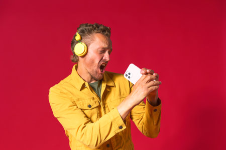 Singing handsome man enjoying his favorite song using phone and wireless headphones wearing jeans yellow jacket isolated on red background. Joyful man sing while recording his voice using smartphone.の写真素材